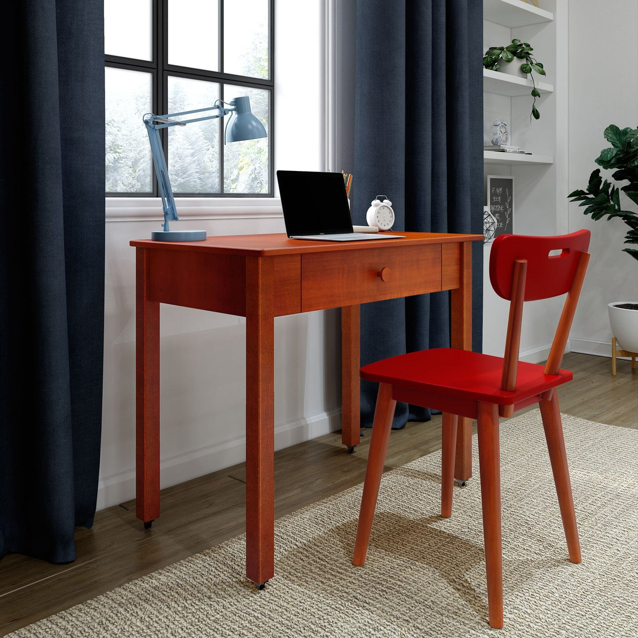 Small wooden desk with drawer, red chair, laptop and blue lamp in modern home office with navy curtains and hardwood floor