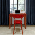 Home office workspace with red chair and wooden desk featuring a laptop, blue desk lamp, pencil holder and alarm clock by a window with dark curtains