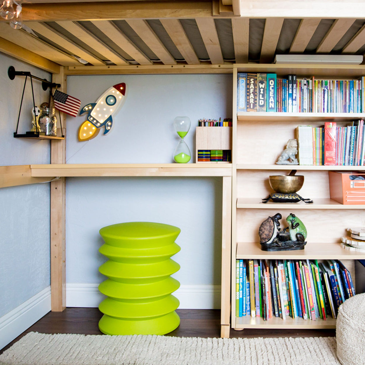 Stylish kids' room with wooden loft bed, decorative rocket, shelves with books, and vibrant green stool.