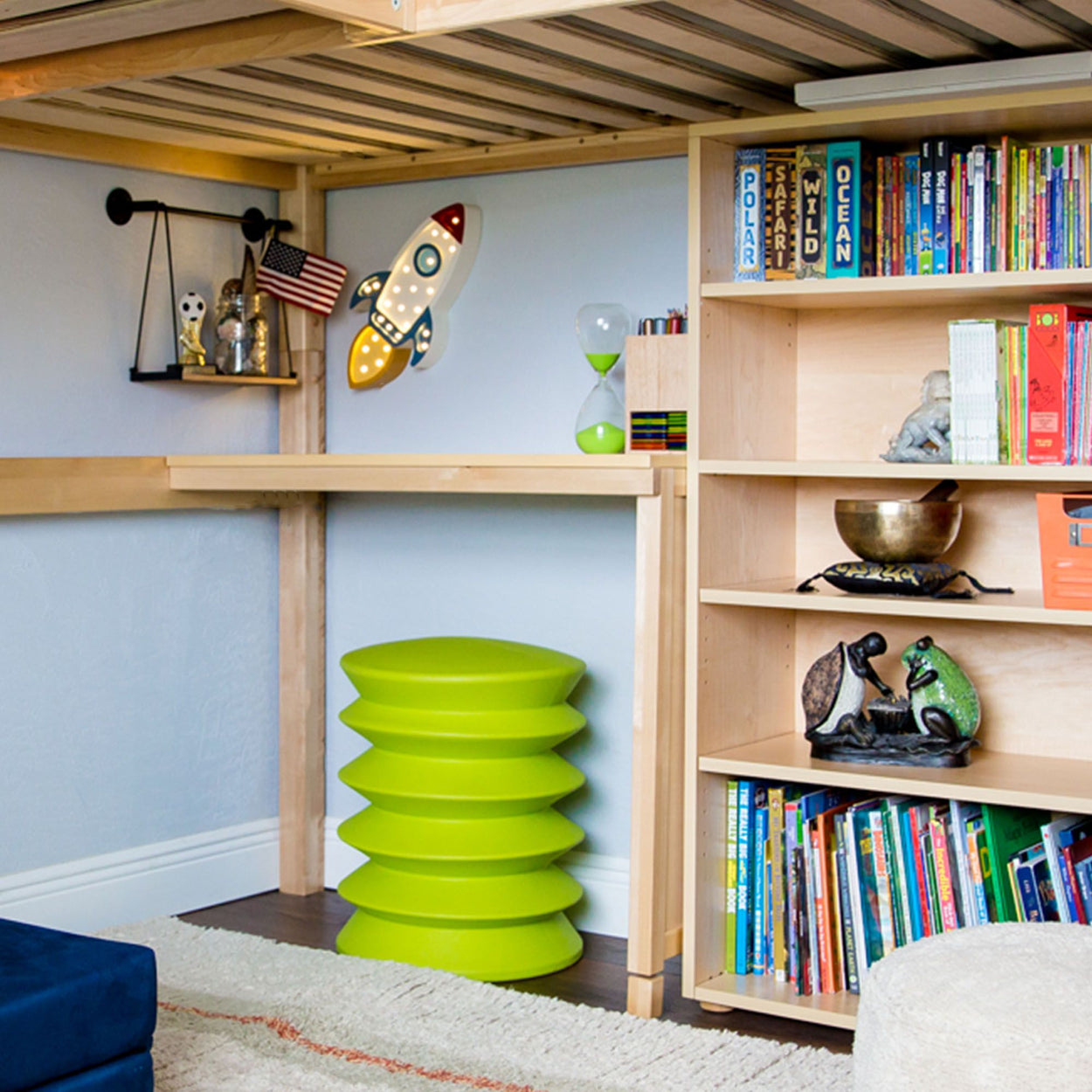 Children's study area with wooden shelves, books, decorative items, and a green stool under a loft bed.