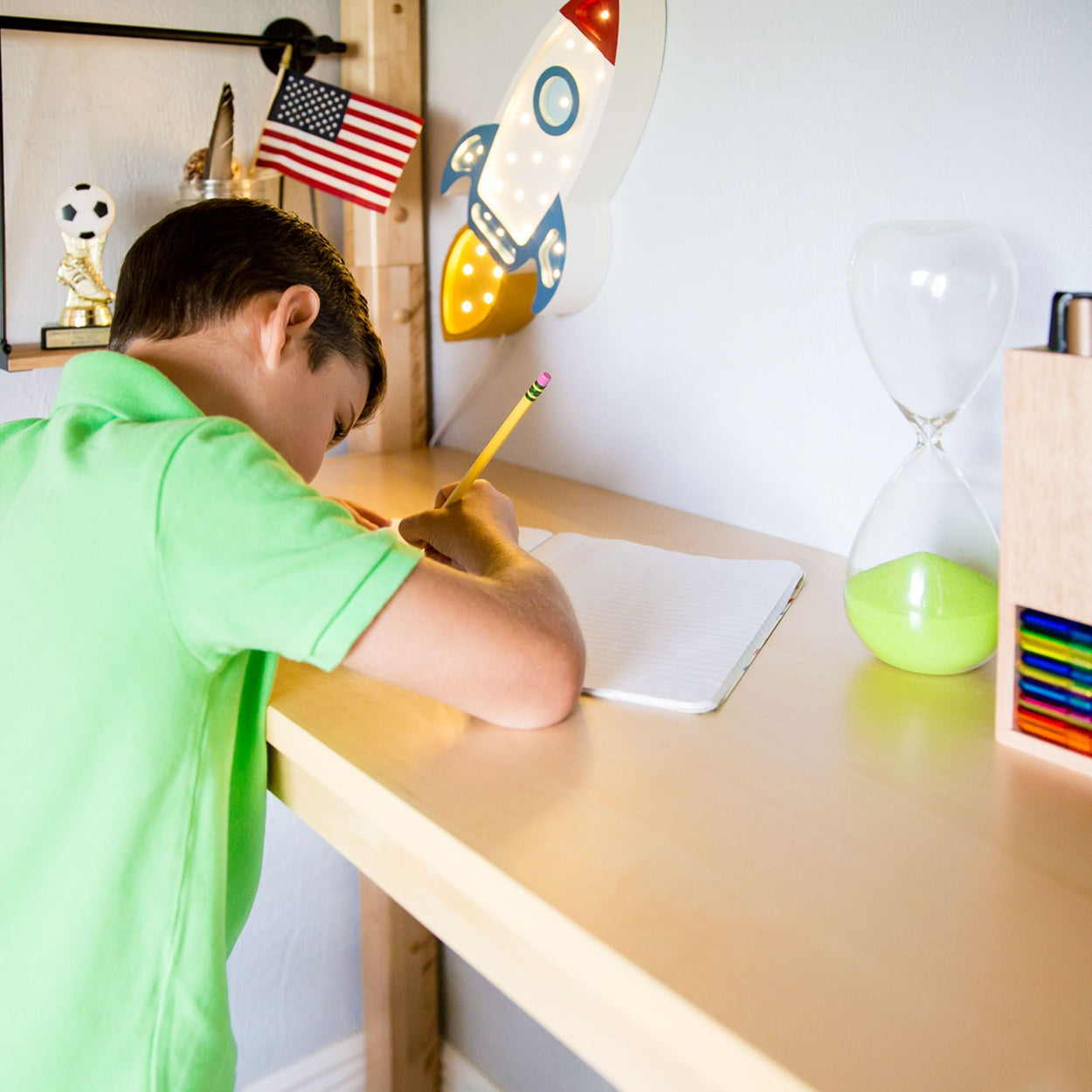 Child writing at a desk with a rocket lamp, American flag, and hourglass, highlighting creative study environment for kids.