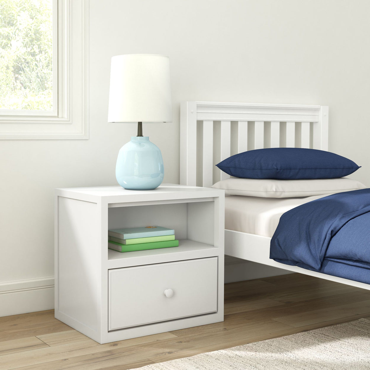 White wooden bedside table with a blue ceramic lamp and books, next to a bed with navy blue bedding in a modern bedroom setting.
