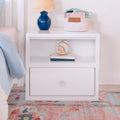 White kids nightstand with blue ceramic lamp, basket of ABC board books and rainbow alarm clock on pastel rug