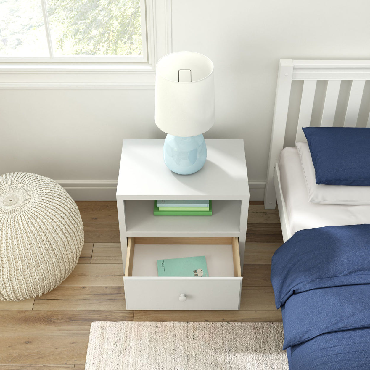 White nightstand with open drawer beside bed, featuring a blue lamp and books, in a bright modern bedroom setting.
