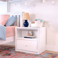 Kids bedroom nightstand with blue ceramic table lamp and woven shade, white bedside table with basket of ABC books, pastel pink and blue bedding, hanging paper lanterns and colorful rug