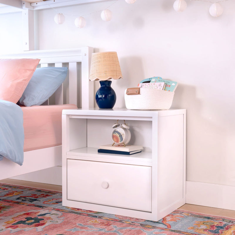 Kids bedroom nightstand with blue ceramic table lamp and woven shade, white bedside table with basket of ABC books, pastel pink and blue bedding, hanging paper lanterns and colorful rug
