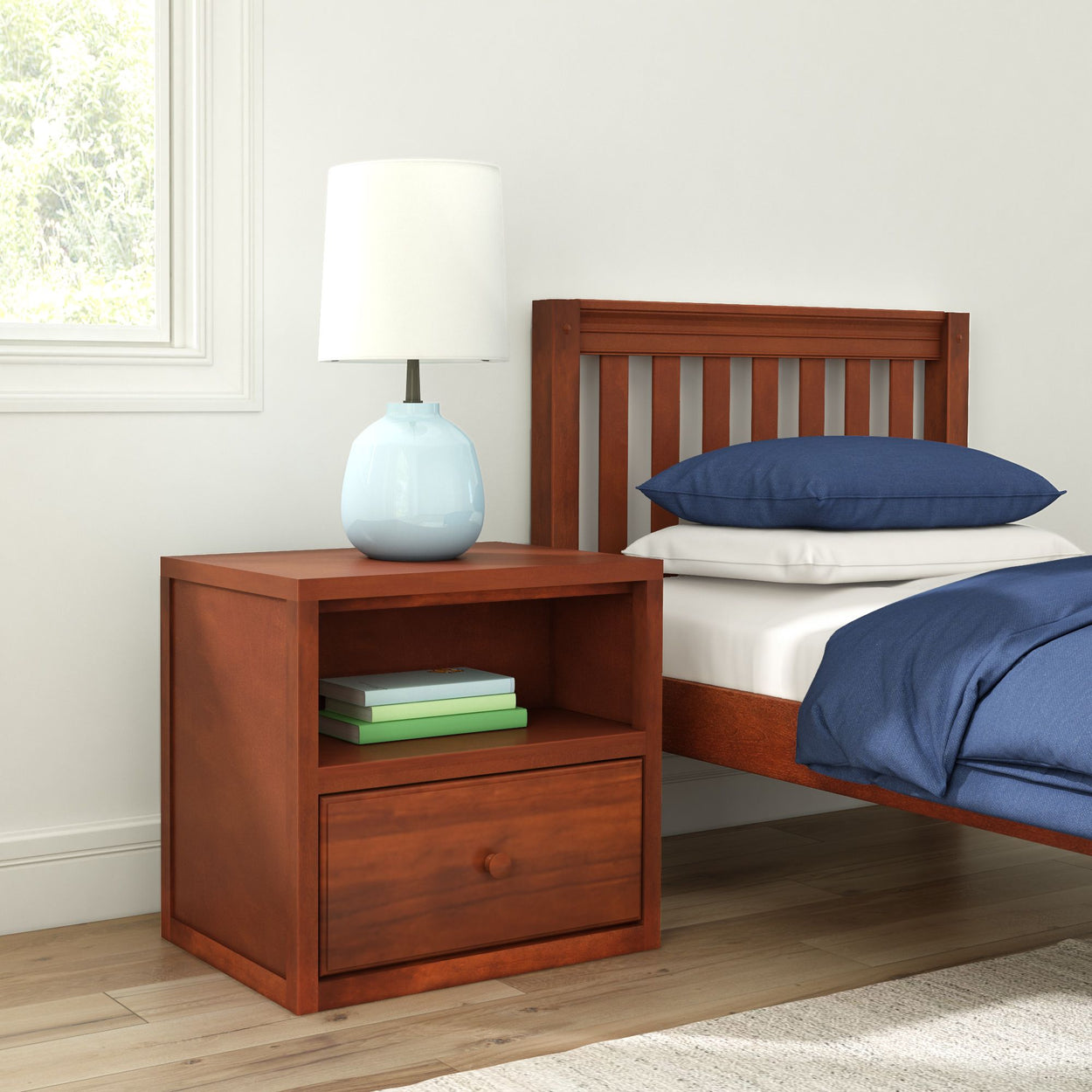 Wooden nightstand with drawer beside bed, featuring a lamp and books.