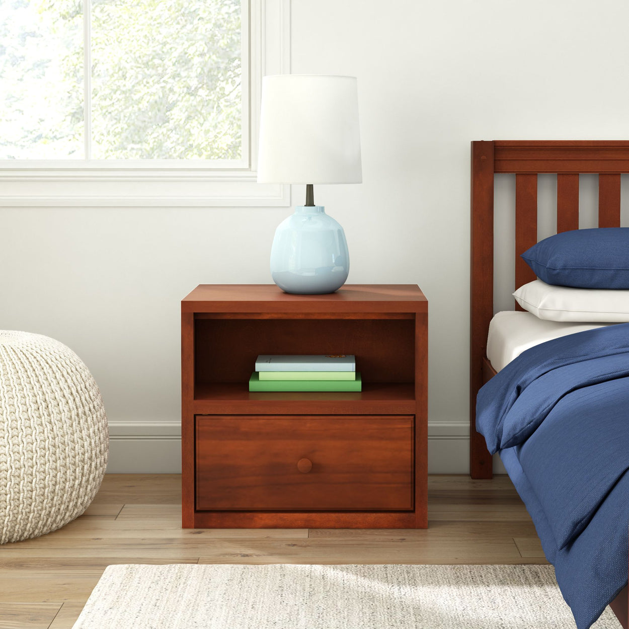 Wooden bedside table with lamp and books next to a bed in a cozy bedroom setting.