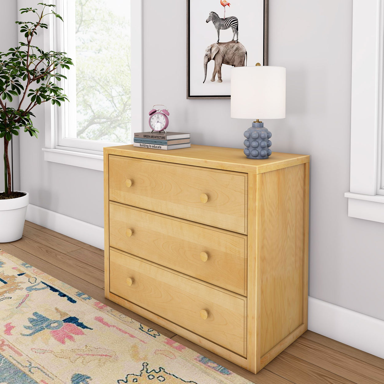 Natural wood dresser with three drawers, topped with a lamp and books, next to a potted plant in a modern bedroom setting.
