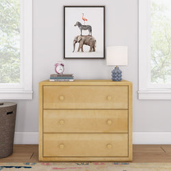 Cream dresser with three drawers, topped with books, a pink clock, and a blue lamp, next to a window, featuring animal art on the wall.