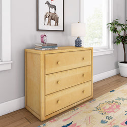Light wood three-drawer dresser in a bright room with a potted plant, decorative books, and a table lamp.