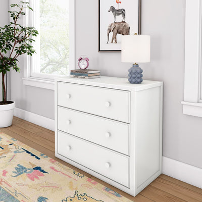 White three-drawer dresser in modern nursery with blue bubble lamp, pink alarm clock on books, framed zebra-elephant-flamingo art, potted plant, pastel area rug and hardwood floor