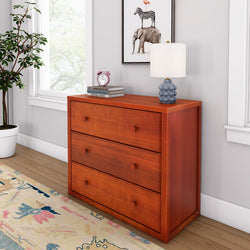 Wooden dresser with three drawers, topped with a lamp, books, and an alarm clock, next to a leafy plant and window in a modern room.