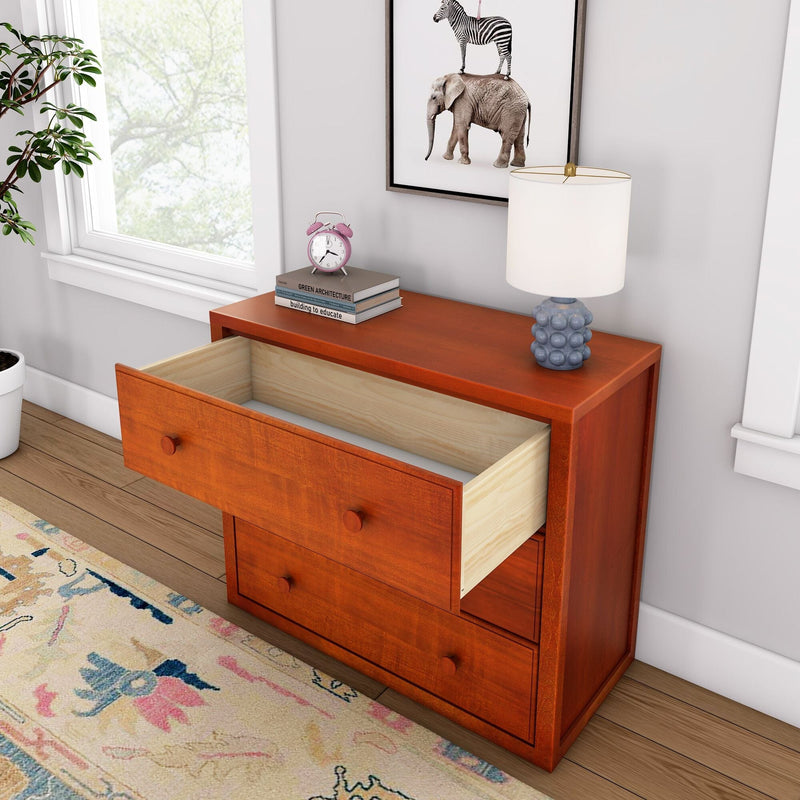 Reddish wooden three-drawer dresser with one open drawer in a light-filled room, styled with a white table lamp, pink alarm clock, stacked books, framed zebra-and-elephant print, potted plant, hardwood floor and colorful rug