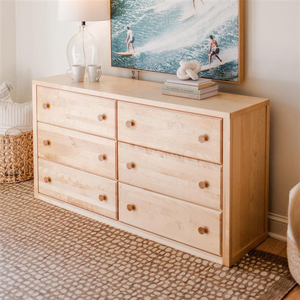 Light maple six-drawer dresser with round wooden knobs in modern coastal bedroom, lamp, surf artwork, woven basket and neutral patterned rug
