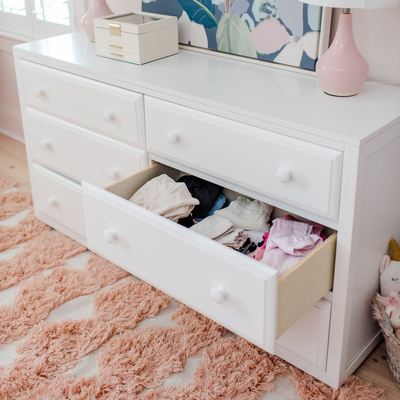 White dresser with open drawer of folded children's clothes, pink table lamp, and plush pink shag rug in a kids' bedroom
