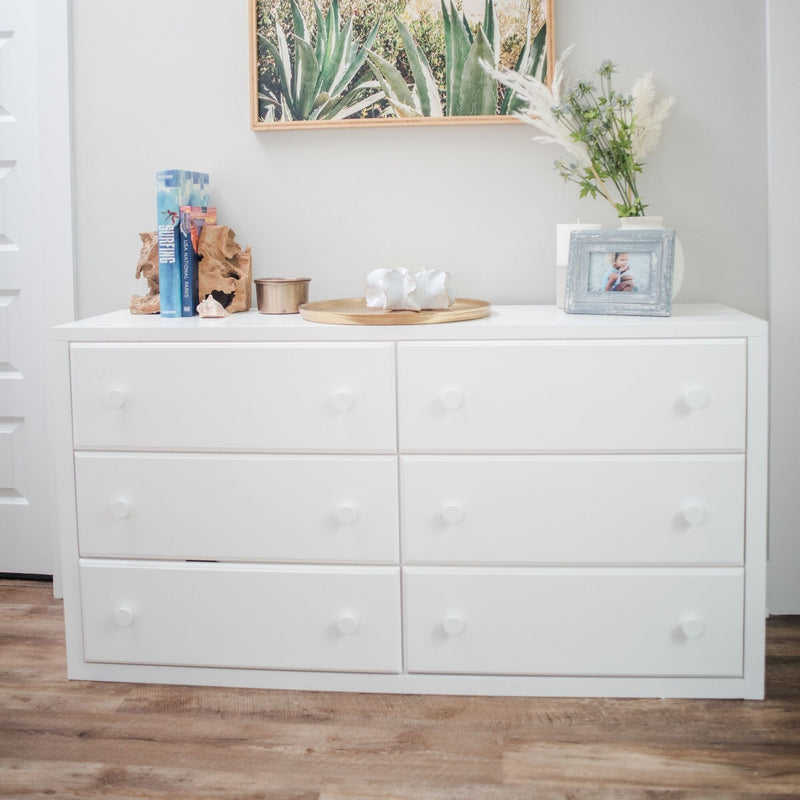 White six-drawer modern dresser in a minimalist bedroom on wood floor, styled with succulent wall art, books and driftwood, a gold tray with seashells, framed photo and vase of pampas grass, bedroom storage and coastal-inspired decor