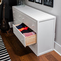 White dresser storage with open drawer showing neatly folded clothes in a modern bedroom with hardwood floor, acoustic guitar and striped rug