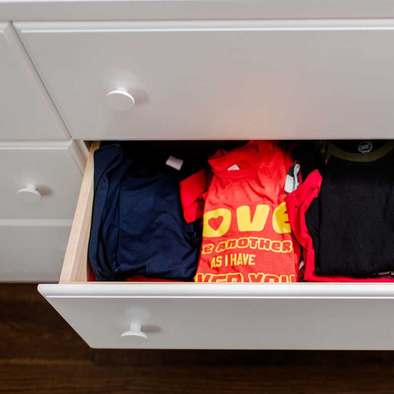 Open white dresser drawer with neatly folded t-shirts — red "LOVE" graphic tee, navy and black shirts; bedroom dresser organization.