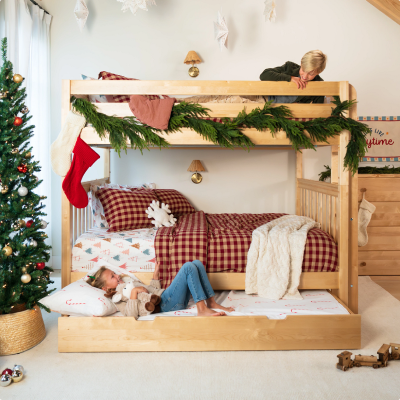 A light natural wood bunk bed with green garland, red and white checkered bedding, and two children playing in a decorated children's bedroom with a Christmas tree.