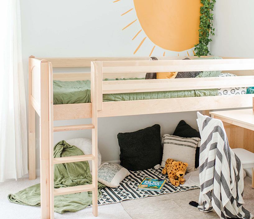 Light wood loft bed with ladder, green bedding, cozy play nook with pillows and stuffed leopard toy, sun wall mural and study desk in children's bedroom