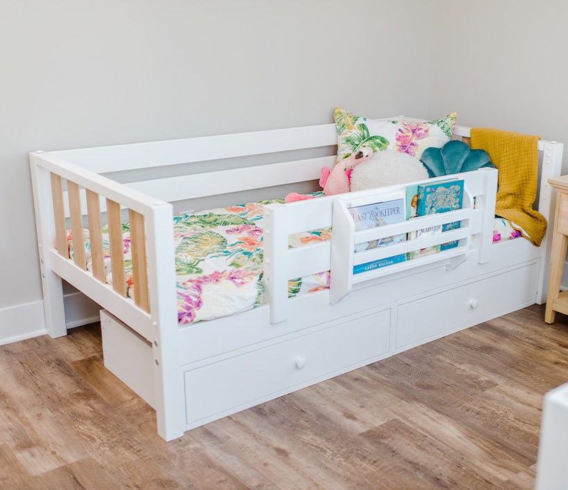 White toddler daybed with safety rail, built-in under-bed storage drawers, colorful floral bedding and plush toys in a modern kids' bedroom on hardwood floor