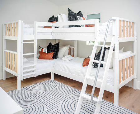 A white and natural two-tone quad corner bunk bed with ladders on each site. The beds have several pillows in multi colors with white bedding to decorate the room. A rug is on the floor with a zen line design.