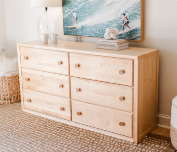 A light natural wood twin-size desk with three drawers, styled next to a wooden bunk bed and a matching wooden chair with a white seat in a bright room.