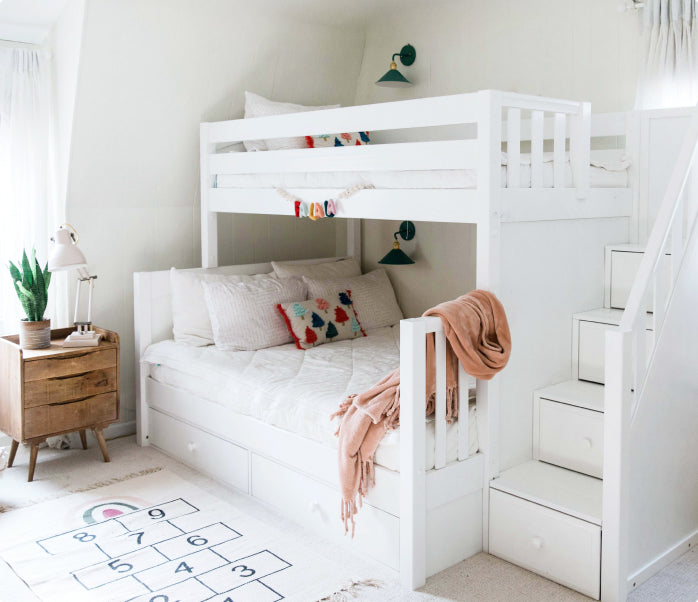 White wooden bunk bed with built-in staircase drawers in a cozy kids' bedroom with trundle bed, hopscotch rug, nightstand and potted plant.