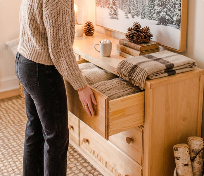 Person opening drawer in cozy winter entryway with wooden dresser, folded knit blankets, plaid throw, pinecone decor, mug of hot chocolate with marshmallows, and snowy forest wall art.