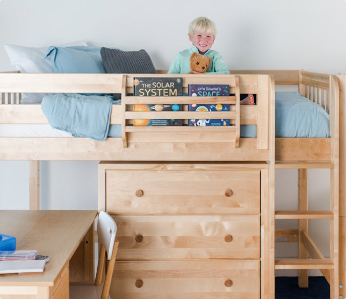Smiling blond child holding a teddy in a light-wood loft bed with ladder, built-in drawers and study desk, blue bedding and space books — kids loft bed with storage and workspace.
