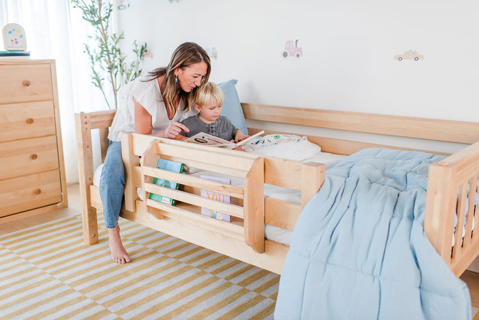 Mother reading to toddler in natural wood toddler bed with built-in book rack, light blue comforter and minimalist kids' bedroom decor