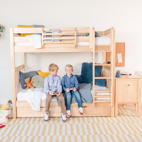 Kids bedroom with a natural wood bunk bed and two boys playing video games on the lower bunk, surrounded by cushions, a teddy bear, desk and corkboard