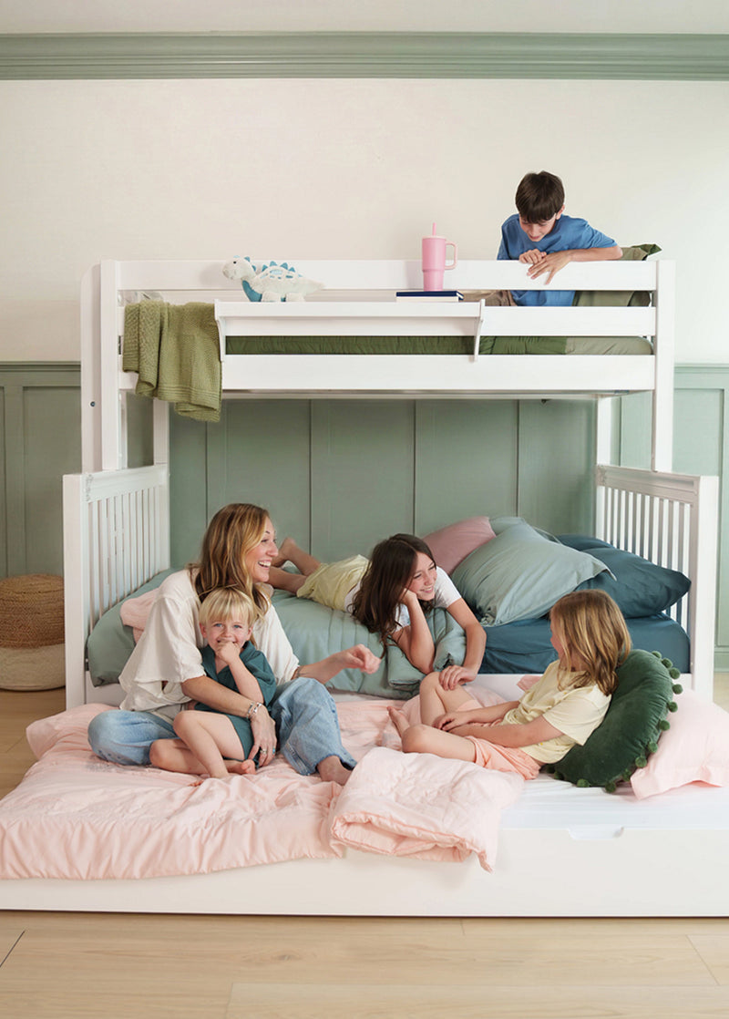 A white wooden bunk bed in a bright bedroom setting, with a family of five interacting on and around the bed. The top bunk has a boy reading, while the bottom bunk features a woman, a young boy, a girl lying down, and another girl sitting up, all surrounded by soft pink and blue bedding.