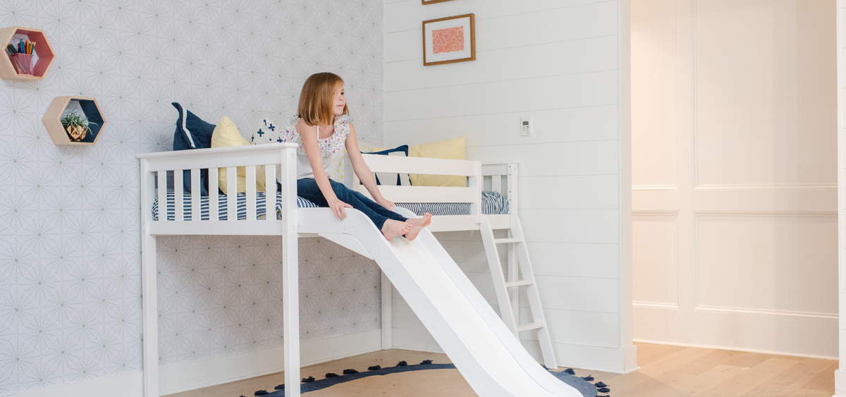 White loft bed with built-in slide in a modern children's bedroom, person sitting on edge, navy and yellow pillows, striped mattress, hexagonal wall shelves and geometric wallpaper
