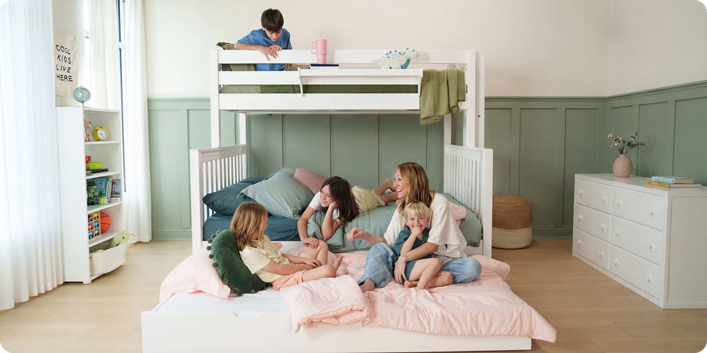 Cozy kids bedroom with white wooden bunk bed and pull-out lower bed, smiling mother and three children playing on pastel pink bedding, green wainscoting, dresser and open toy shelf — modern family-friendly bunk bed room décor.