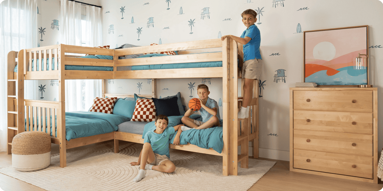 Wooden L-shaped kids bunk bed with teal bedding and checkered pillows in a coastal-themed bedroom with palm wallpaper, wooden dresser, and three boys.