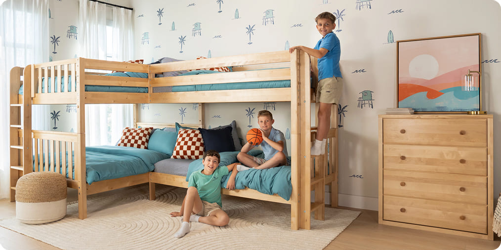 Three boys playing on a natural solid-wood L-shaped bunk bed with turquoise bedding, checker pillows and coastal palm wallpaper in a modern kids' bedroom with light-wood dresser.