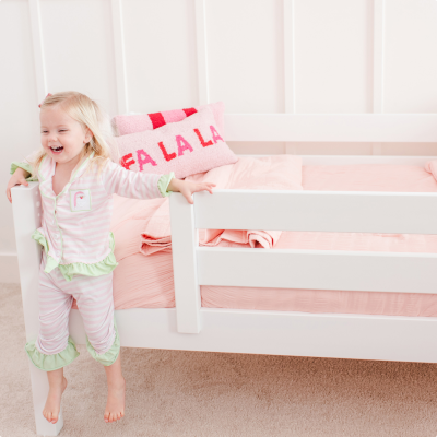 A young girl wearing a pink and white striped pajama set stands next to a white toddler bed with pink bedding and pillows in a bright nursery setting.