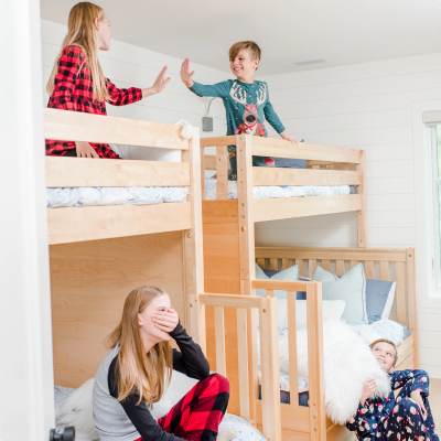 A wooden bunk bed with a built-in climbing ladder in a bright bedroom, with four children playing and interacting on and around the beds.