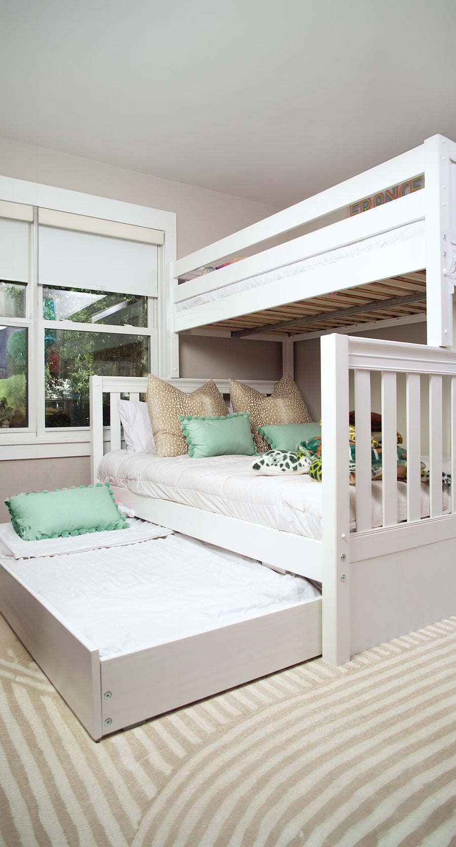 A natural finish twin XL over Queen bunk bed with trundle bed pulled out. The room has white, beige, and light green pillows to decorate the bed along with white bedding.