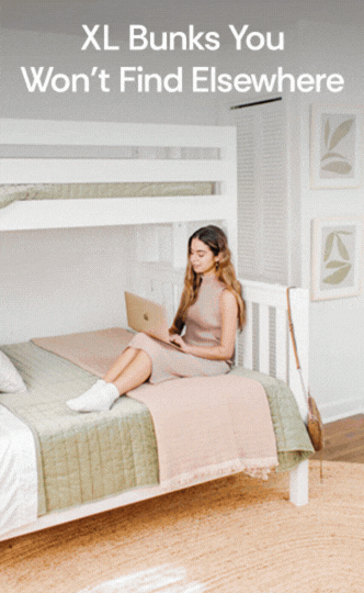 Person using a laptop on the lower bunk of a white XL extra-long wooden bunk bed in a bright modern bedroom with green and pink bedding, rattan rug and framed wall art, ideal space-saving bunk bed for teen bedrooms and dorm rooms