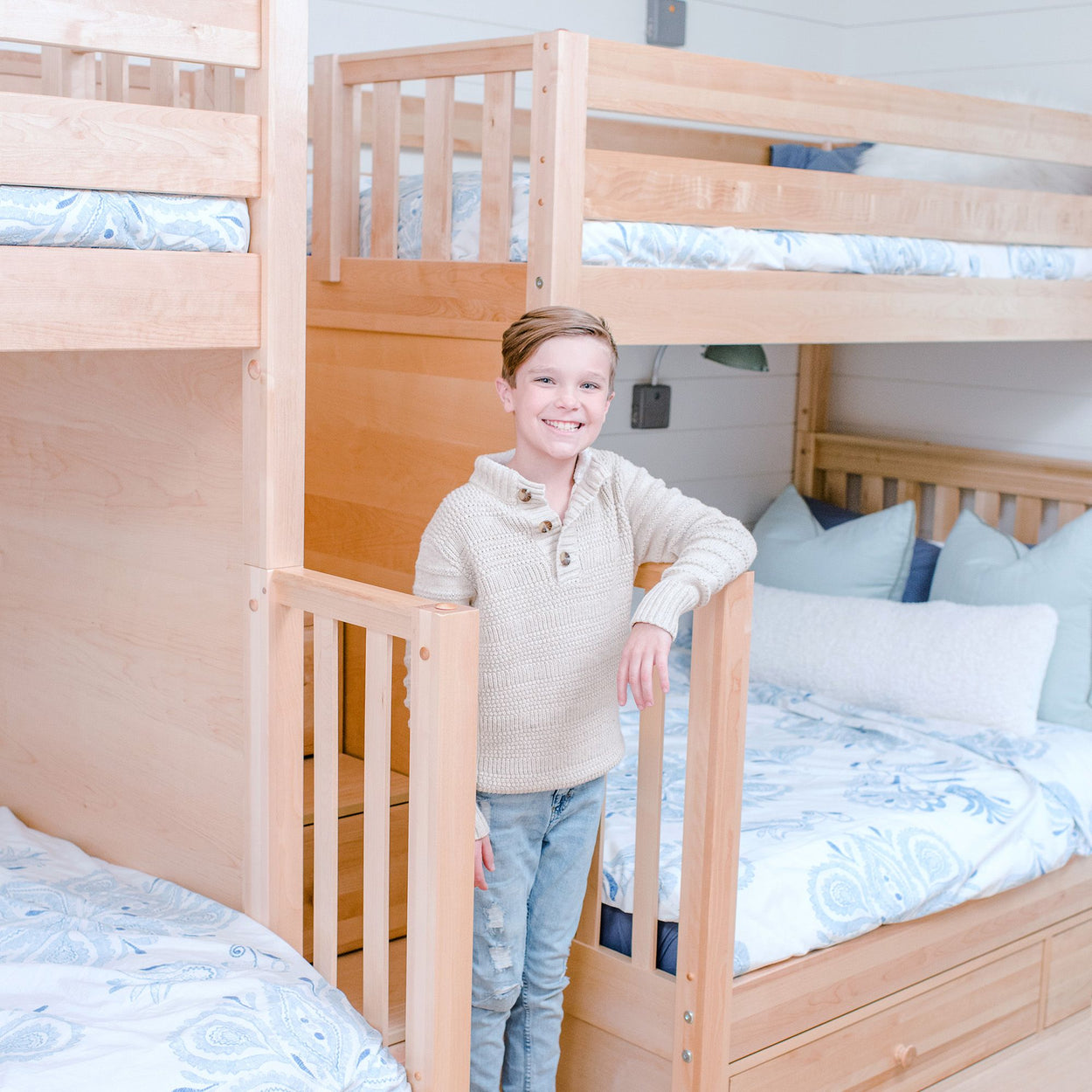 Boy smiling in cozy bedroom with wooden bunk beds and decorative pillows.