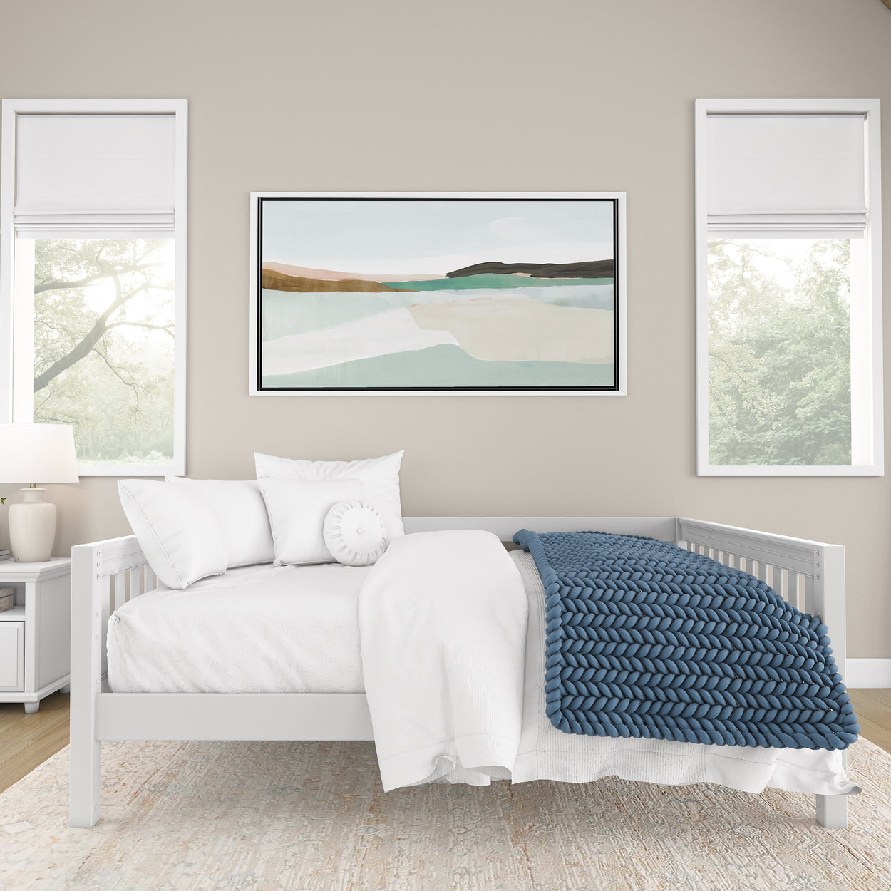 Minimalist bedroom with a white bed, blue knitted blanket, abstract landscape artwork, and large windows for natural light.