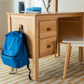 Light wood home study desk with blue backpack hanging, pencil cup and lunchbox on top, white chair on striped rug