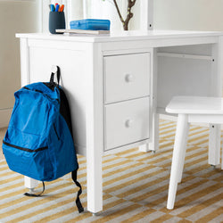 White kids' study desk with storage drawers and chair, blue backpack hanging, pen cup and blue lunchbox on top, yellow-striped rug