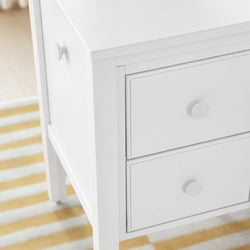 White wooden dresser bedside table close-up with round knobs and drawers on striped rug, modern nursery storage furniture