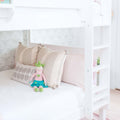 White wooden bunk bed with ladder, pastel pink and beige pillows, patterned wallpaper and knitted doll in a bright children's room