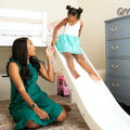 Mother and daughter playing on a white indoor slide attached to a bunk bed in a bright kids' bedroom, playtime scene with toy car and dresser