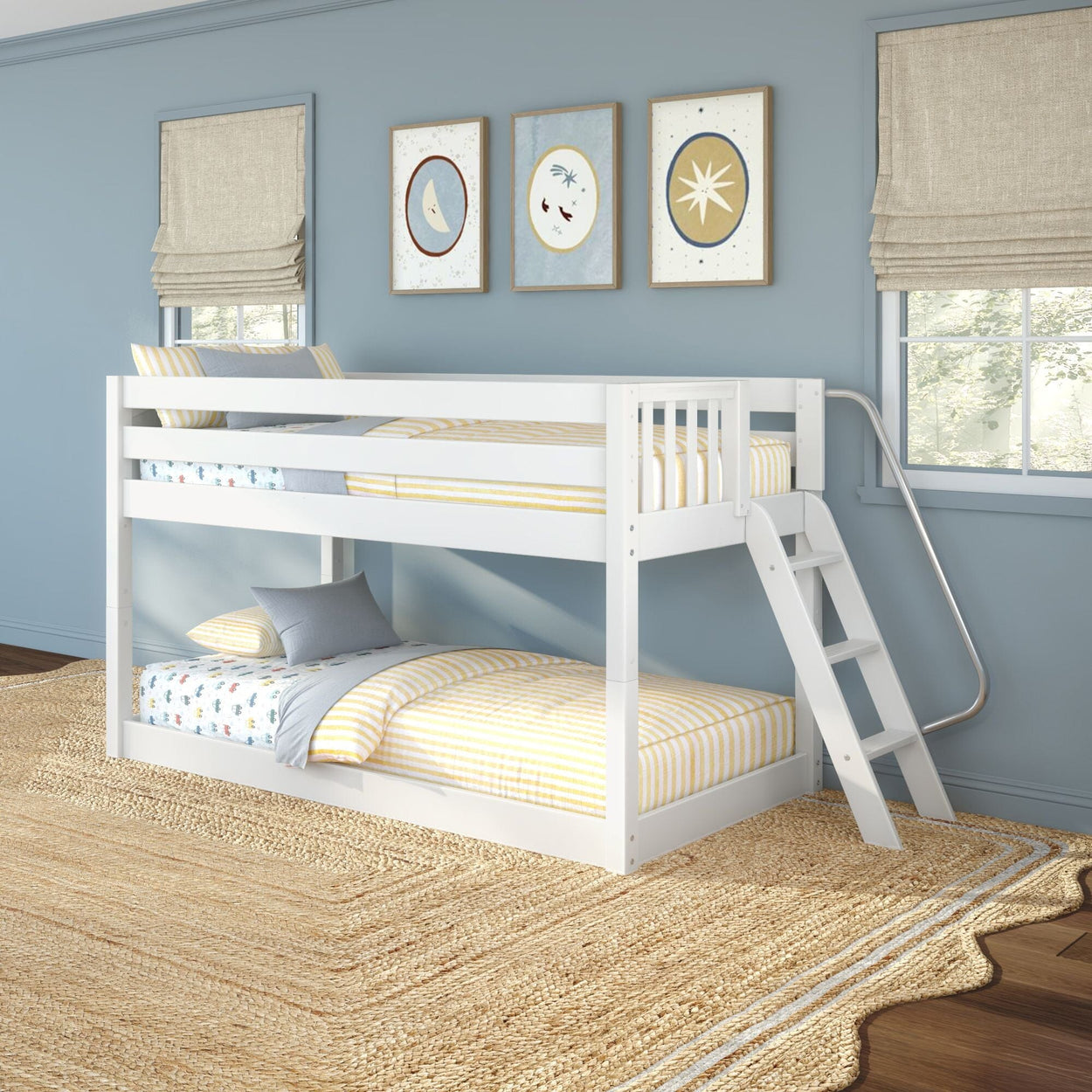 White wooden twin-over-twin bunk bed with ladder in children's blue bedroom, yellow-striped bedding, moon and star wall art, beige roman shades and natural jute rug
